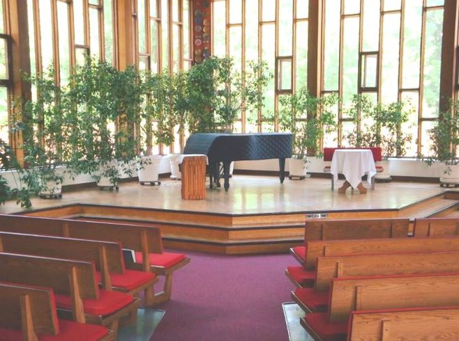 A view of the front of Worship Hall. Our baby grand piano appears on the dais, next to the pulpit, in front of the wall of windows that fills our Sanctuary with natural light. There is burgundy carpet in the aisles and the wooden pews have red cushions on them. There are lots of potted plants in front of the windows.