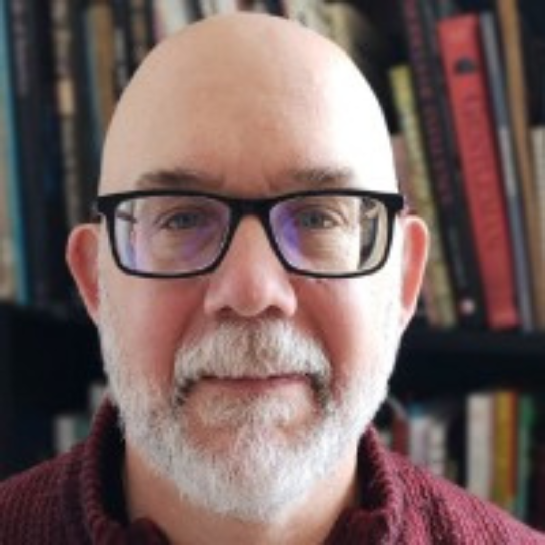 A headshot of Guy Belleperche - a middle-aged white man with a white beard, wearing a burgundy sweater and standing in front of a book case. He is a lay chaplain and a member of First Unitarian Congregation of Ottawa.