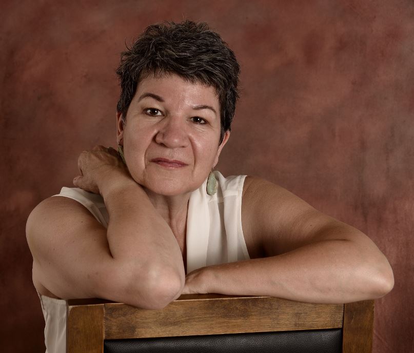 A headshot of Lia Boulay, a middle-aged woman in a white tank top and big earrings. She has a pixie haircut and is sitting backwards in a wooden chair. She is a lay chaplain and a member of First Unitarian Congregation of Ottawa.