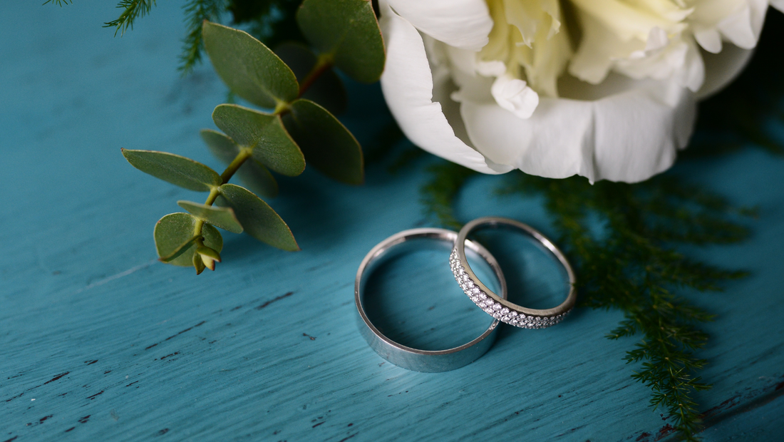 A stock photo of two silver wedding rings next to a delicate bouquet comprised of a white peony and some greenery. They are grouped together on a teal, wooden tabletop.